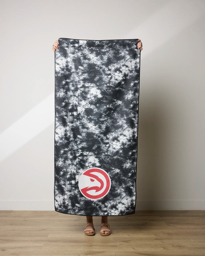 A person holds up the Hawks Flash beach towel by NBALAB x Geometry, featuring a black and white tie-dye design and Atlanta Hawks logo. Only their hands, feet, and sandals are visible. The quick-drying towel rests on an indoor wooden floor.
