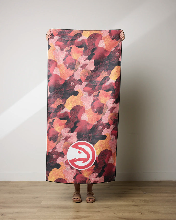 A person stands indoors holding the Hawks Camo NBALAB x Geometry beach towel, featuring a red, pink, and yellow camouflage pattern with a white Atlanta Hawks logo; only their hands and feet are visible.