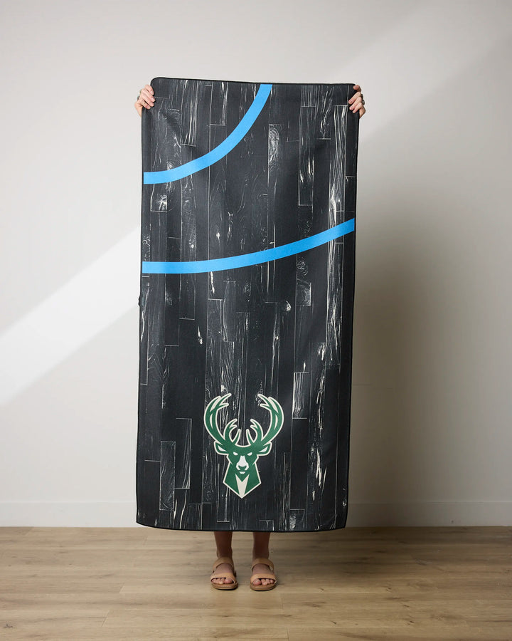 A person stands on a wooden floor holding up the NBALAB x Geometry Bucks Zone beach towel with blue curves and a green Milwaukee Bucks deer logo, made from recycled materials, covering all but their hands, feet, and sandals.