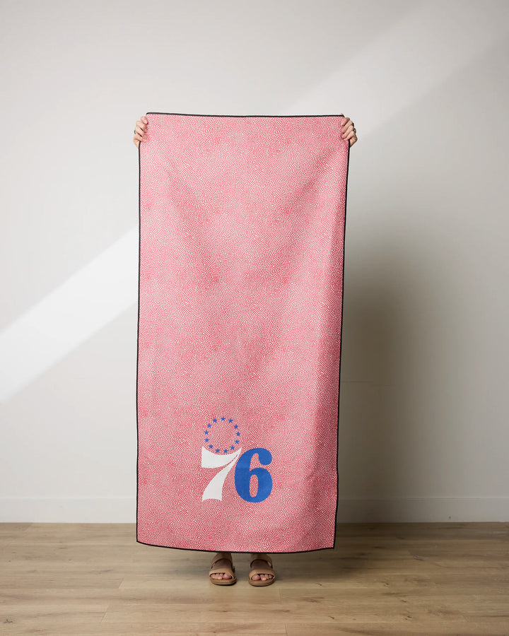 A person stands on a wooden floor holding up the NBALAB x Geometry 76ers Grip towel, a rectangular, pink-patterned, quick-drying beach towel featuring a blue and white "76" and a ring of blue stars in one corner, hiding their head and most of their body.