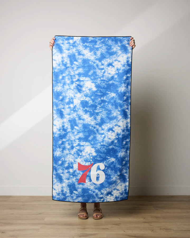 A person holds up the NBALAB x Geometry 76ers Flash beach towel, featuring a blue and white design with the number 76 in red and white on one corner, standing on a light wood floor against a plain wall.