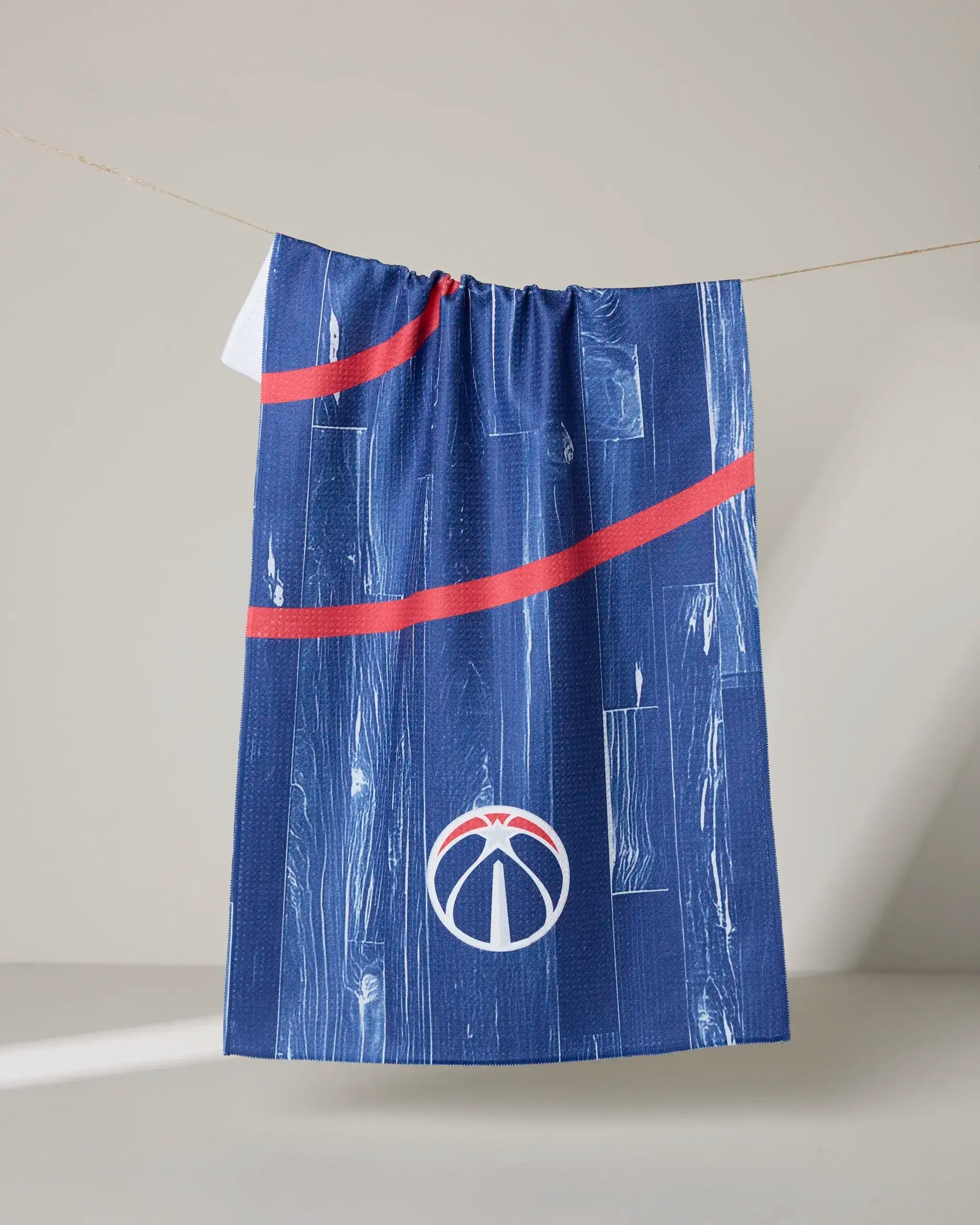 A blue "Wizards Zone" tea towel by NBALAB x Geometry, featuring a wood grain pattern, red diagonal lines, and the white Washington Wizards logo, hangs on a clothesline against a neutral background.