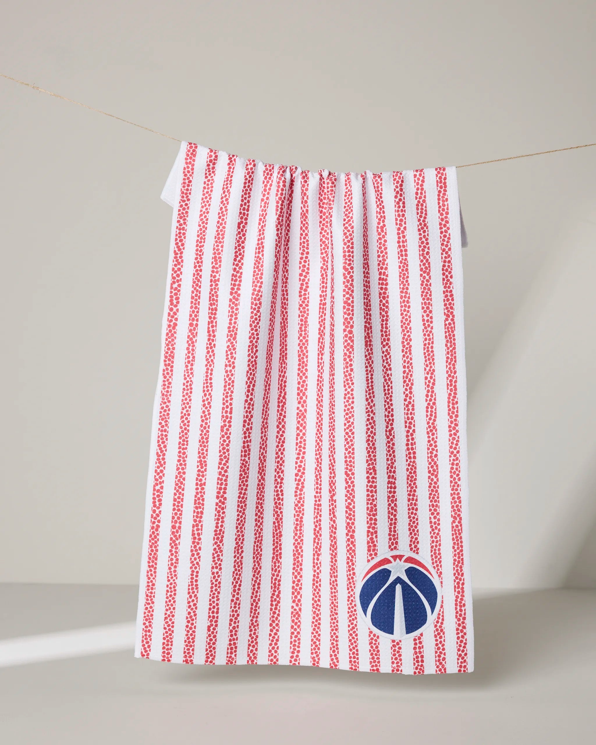 The NBALAB x Geometry Wizards Grip Stripe kitchen towel, featuring vertical red stripes and a blue basketball logo in the bottom right corner, hangs on a clothesline against a neutral background.