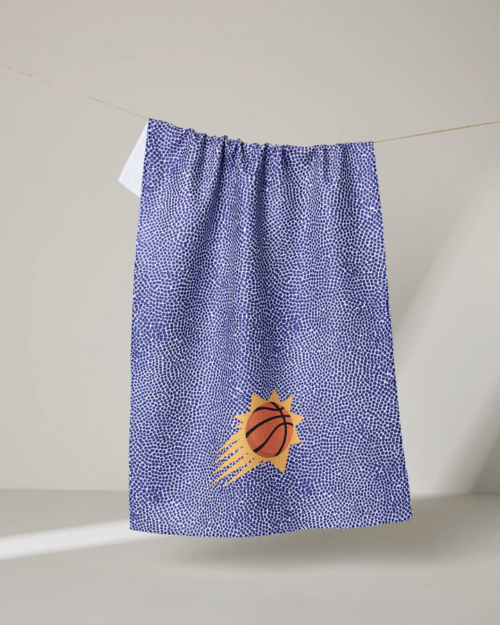 A blue and white speckled NBALAB x Geometry Suns Grip tea towel hangs on a line, featuring a basketball graphic with orange motion lines and a yellow sunburst on the lower half.