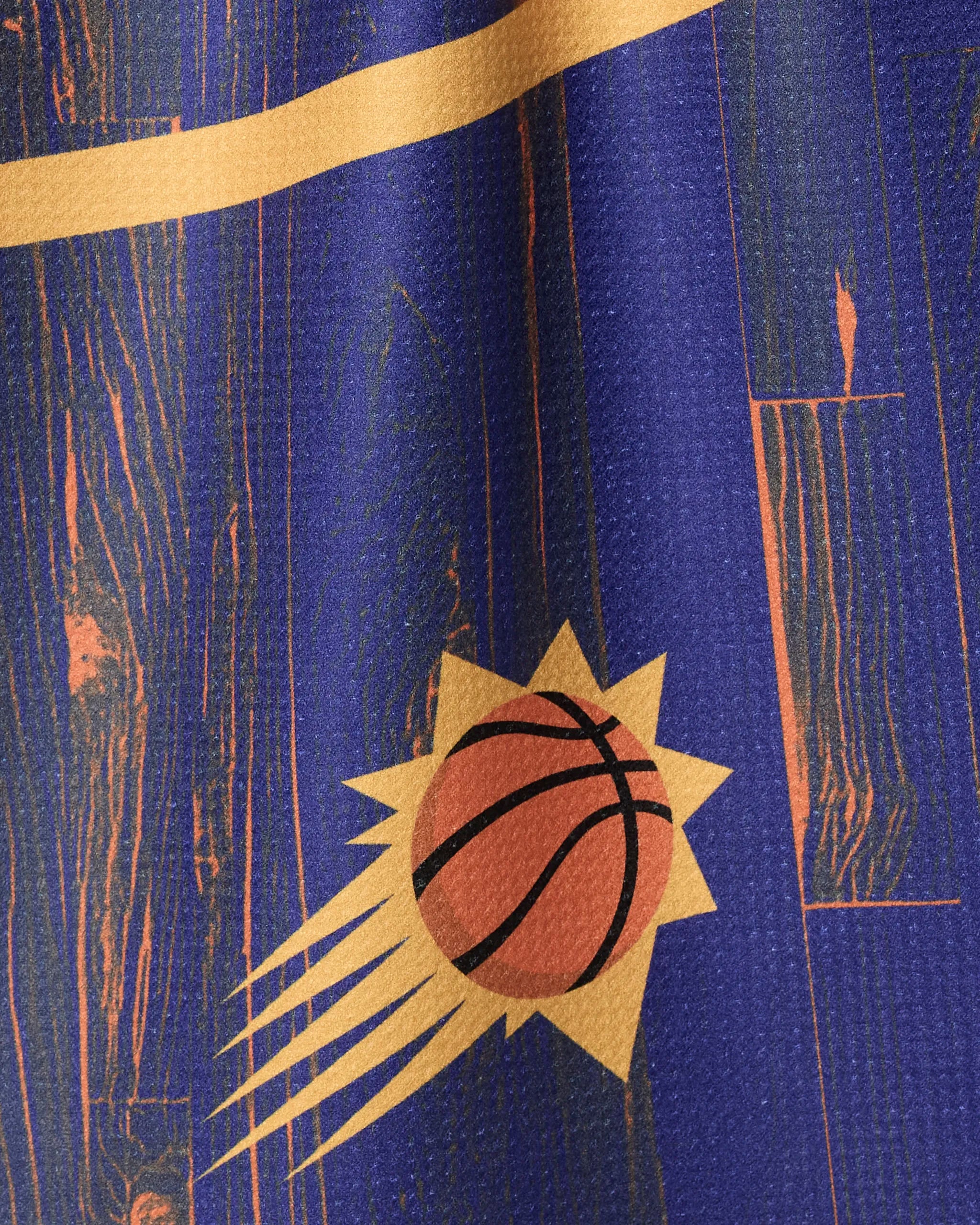 A close-up of the Suns Zone kitchen towel by NBALAB x Geometry, featuring a blue and orange pattern with a stylized basketball and sun rays inspired by the Phoenix Suns logo, plus a woodgrain texture and diagonal orange stripe.