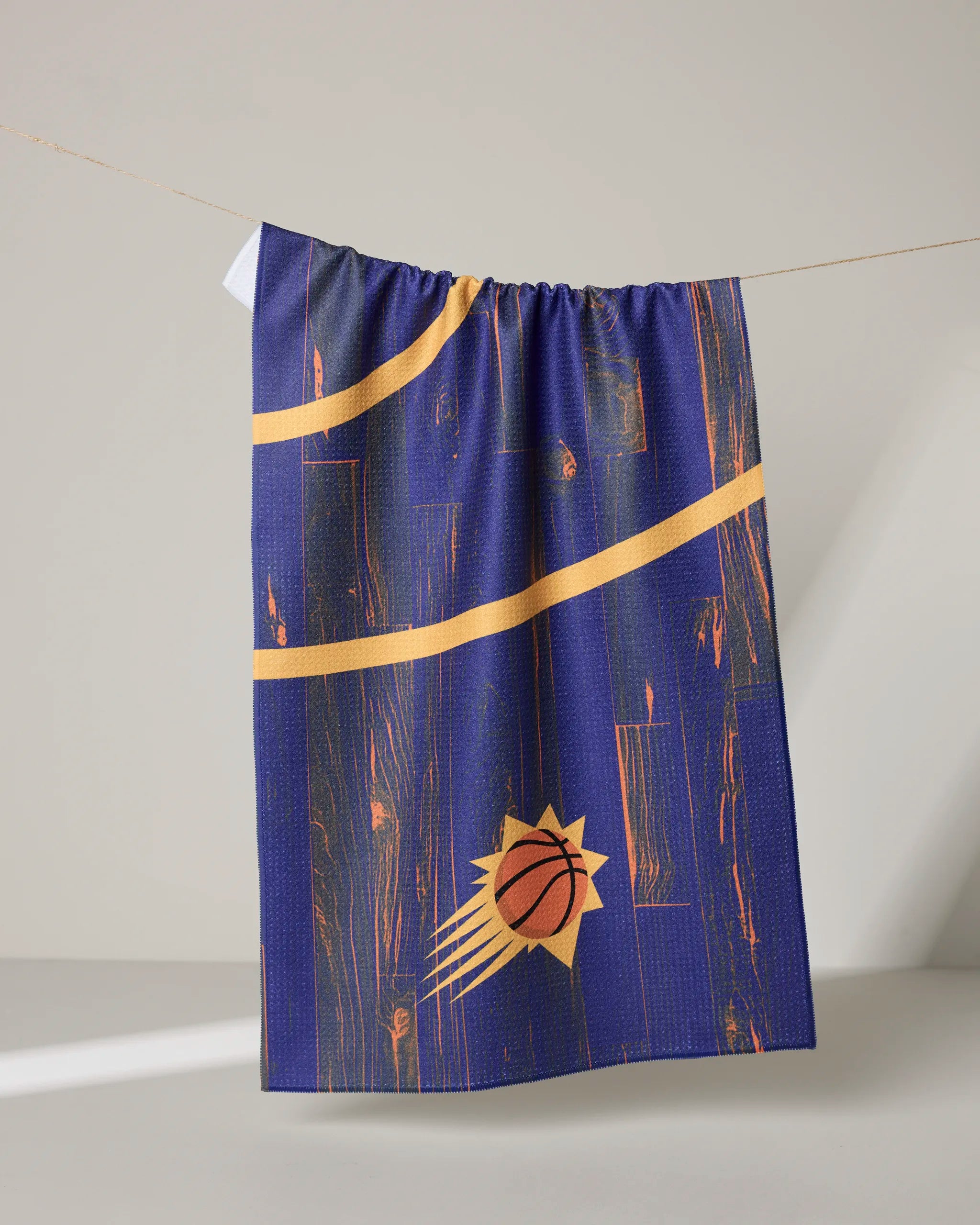 The "Suns Zone" tea towel by NBALAB x Geometry features yellow curved lines and a basketball sunburst logo on blue, hanging against a neutral background—an officially licensed NBA design that brings sporty flair to any space.