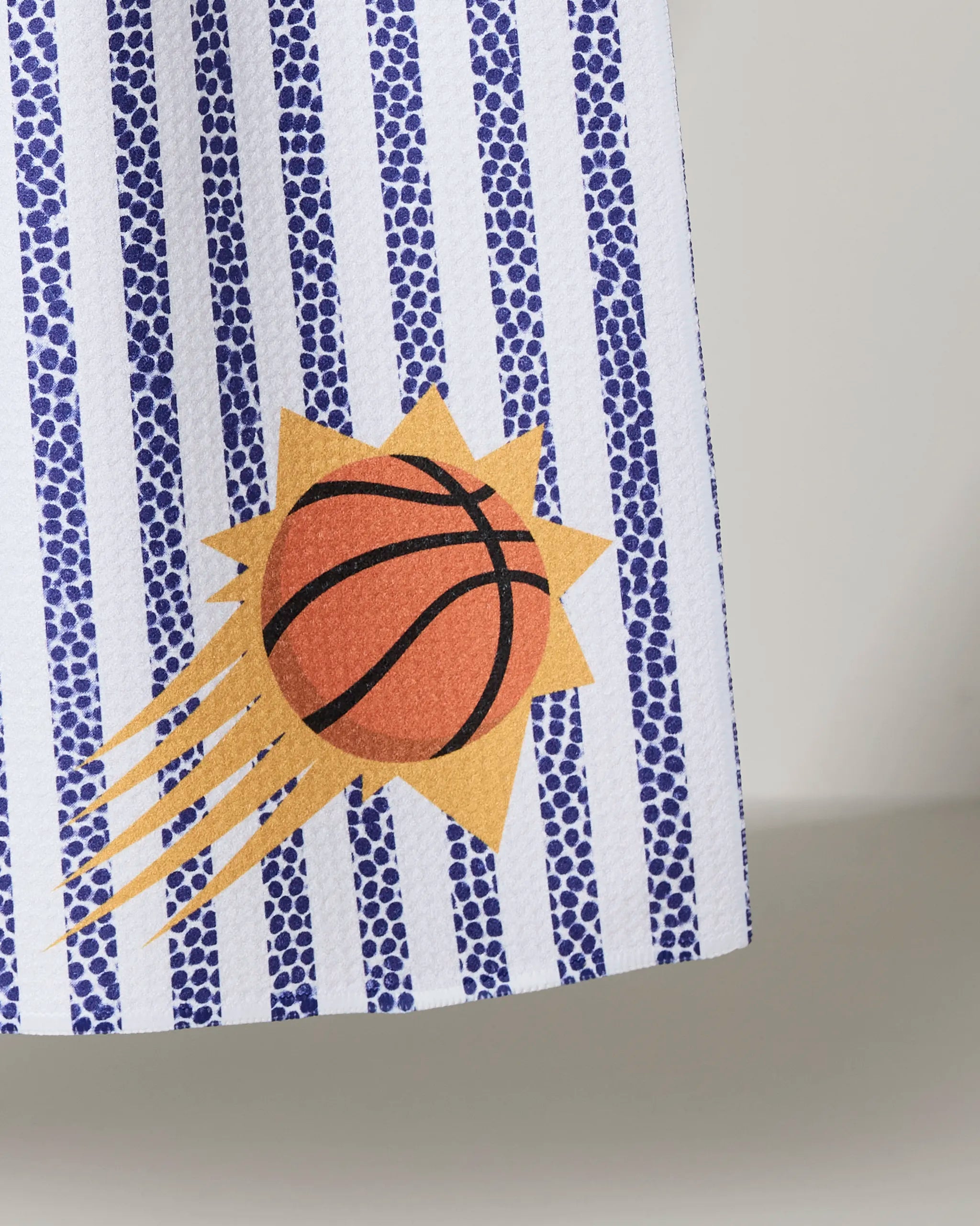 A close-up of the Suns Grip Stripe kitchen towel by NBALAB x Geometry, featuring blue dotted vertical stripes and a graphic orange basketball with motion lines over a yellow sunburst.