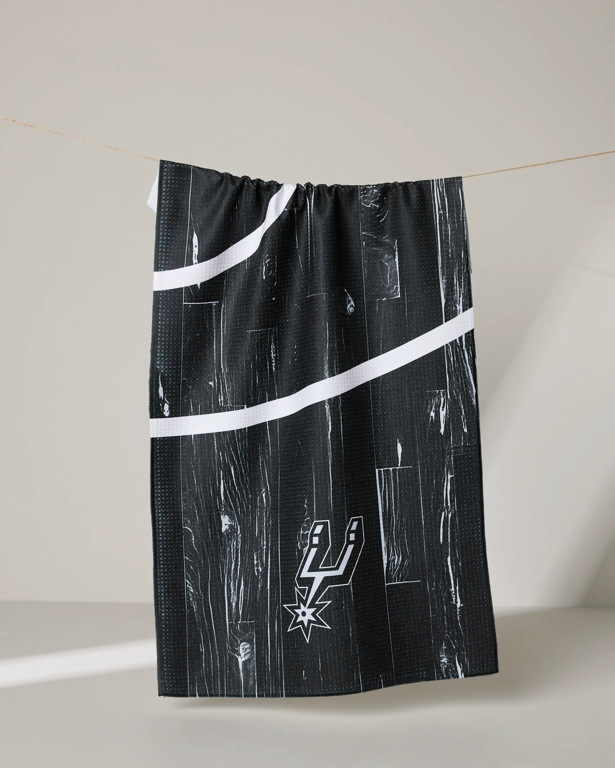 The Spurs Zone tea towel by NBALAB x Geometry features a black recycled design with a wood grain pattern and white lines, hanging on a string. The San Antonio Spurs logo appears in the lower right corner.