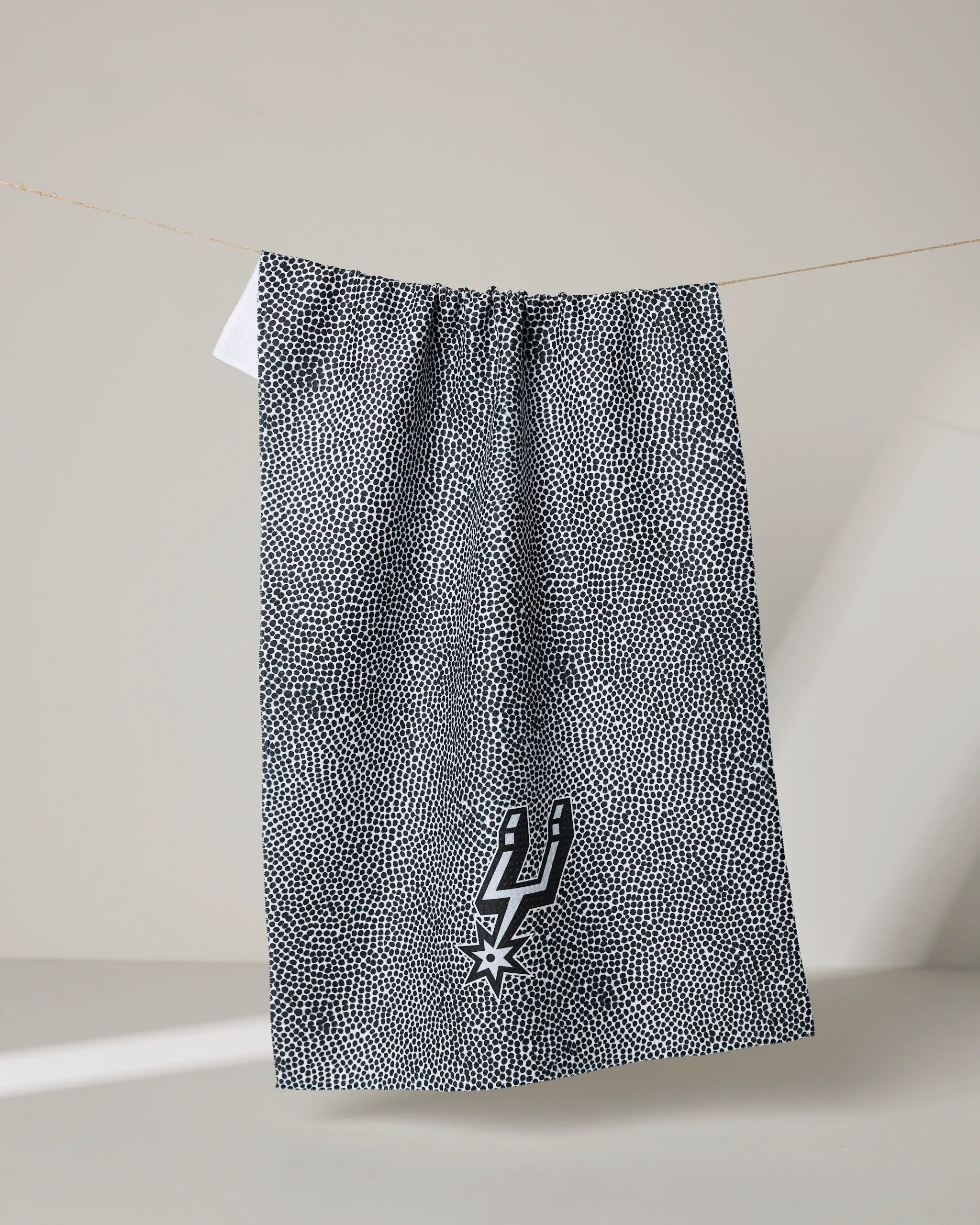 A black and white dotted Spurs Grip tea towel by NBALAB x Geometry hangs on a line, featuring the San Antonio Spurs logo near the bottom against a plain beige background.