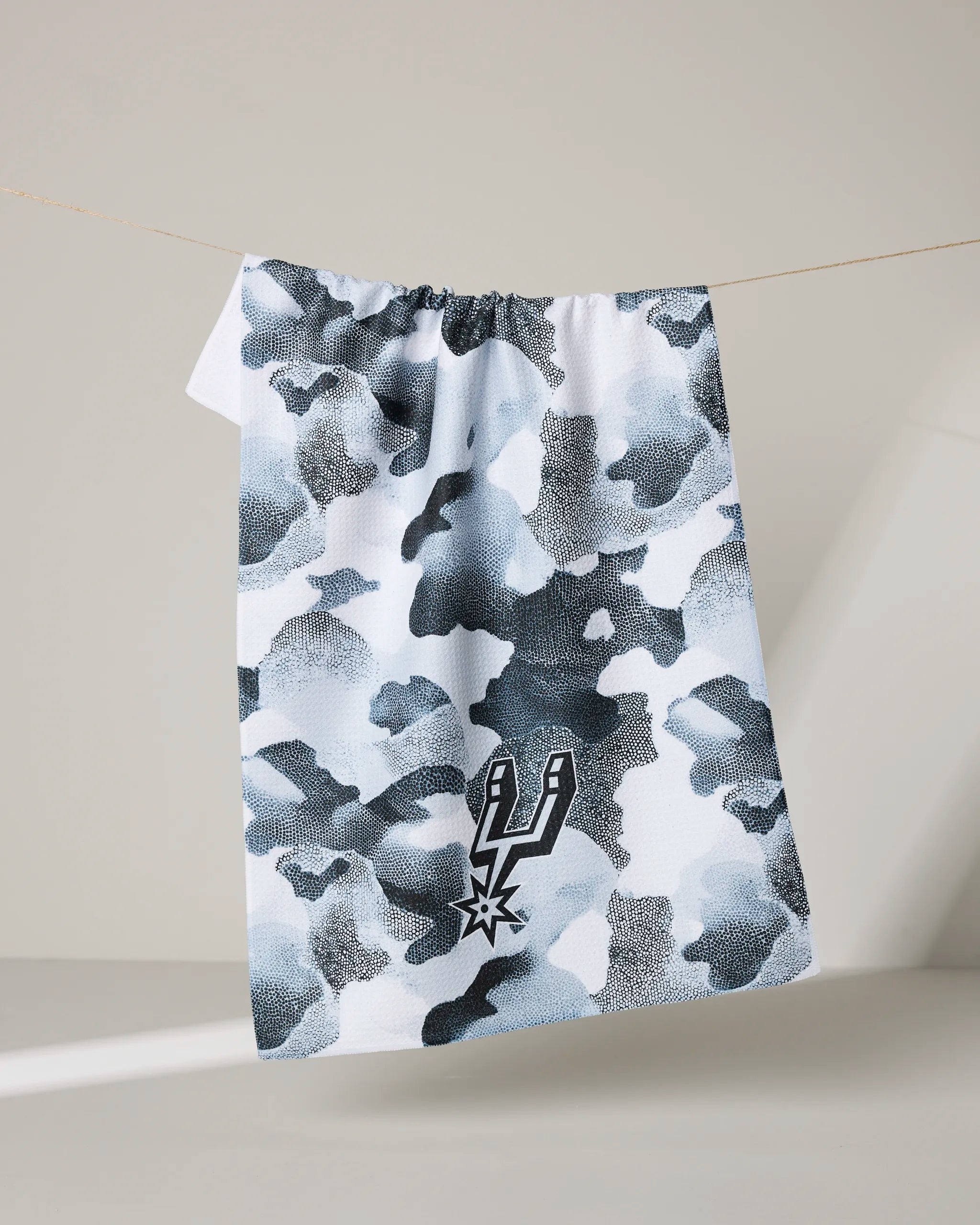 The NBALAB x Geometry Spurs Camo tea towel, with a white and gray camouflage pattern and San Antonio Spurs logo, hangs on a clothesline against a minimalist, light background.