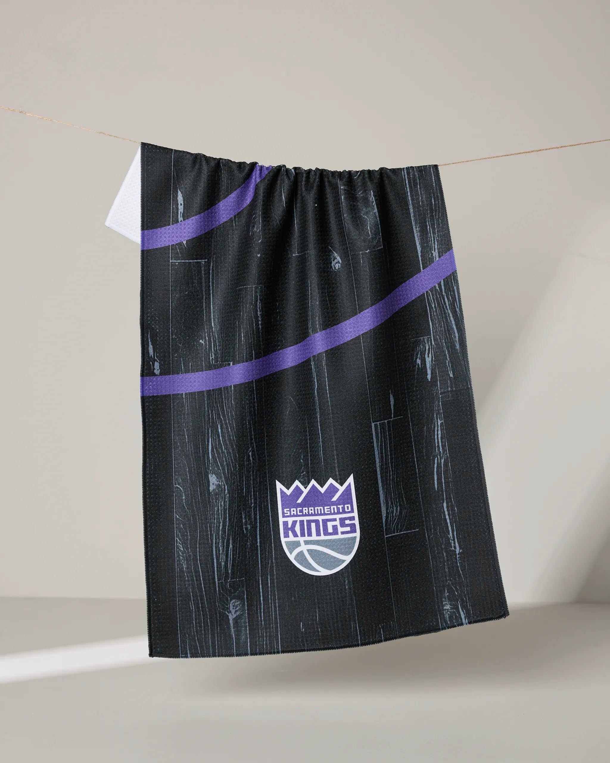 A black Kings Zone tea towel from NBALAB x Geometry with a purple stripe and woodgrain pattern hangs on a line, featuring the Sacramento Kings logo at the bottom center. This sustainable kitchen towel is part of their collection.