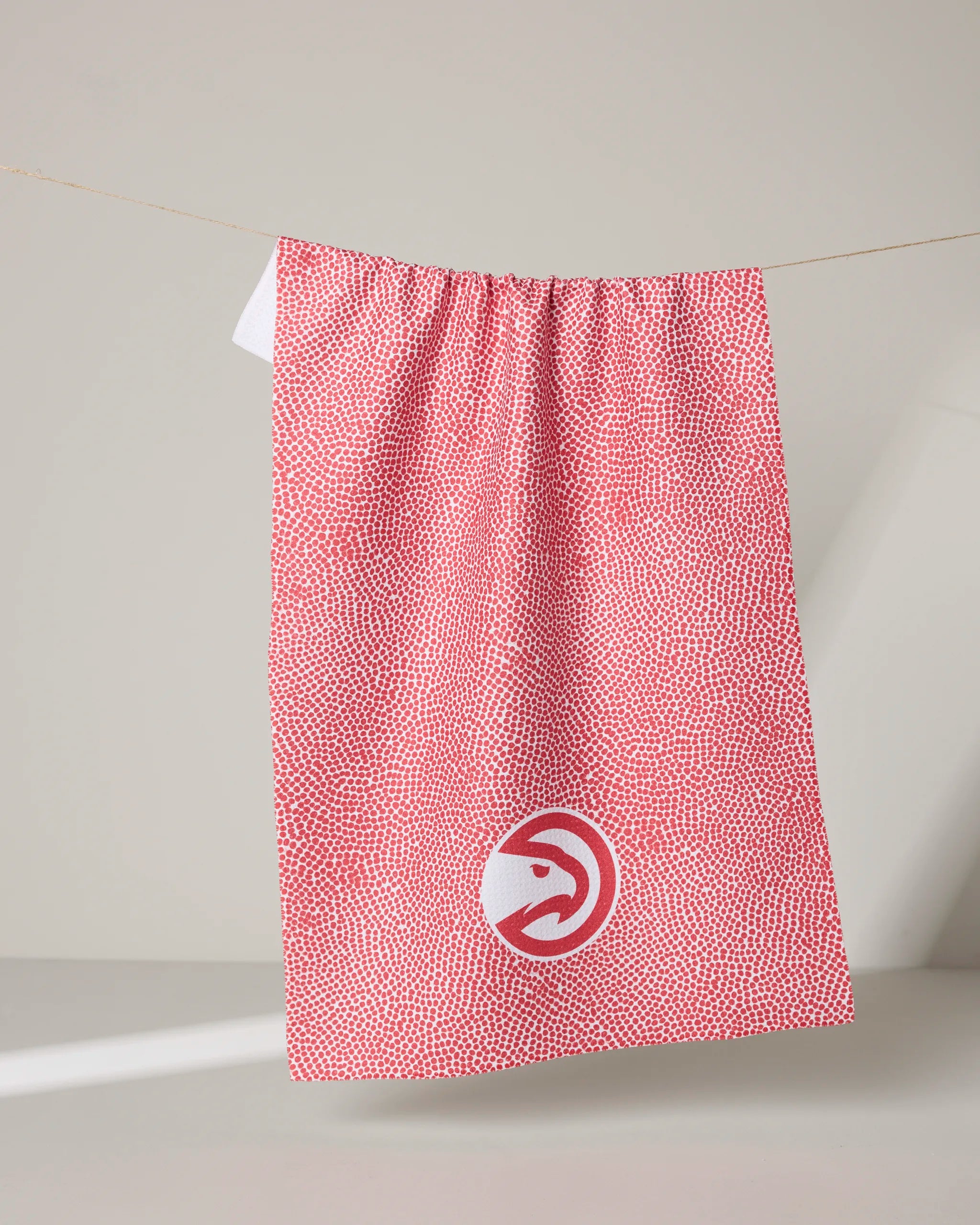 A red and white tea towel called "Hawks Grip" from the NBALAB x Geometry collection, featuring the Atlanta Hawks logo, hangs on a string against a plain background—perfect for stylish NBA fans.