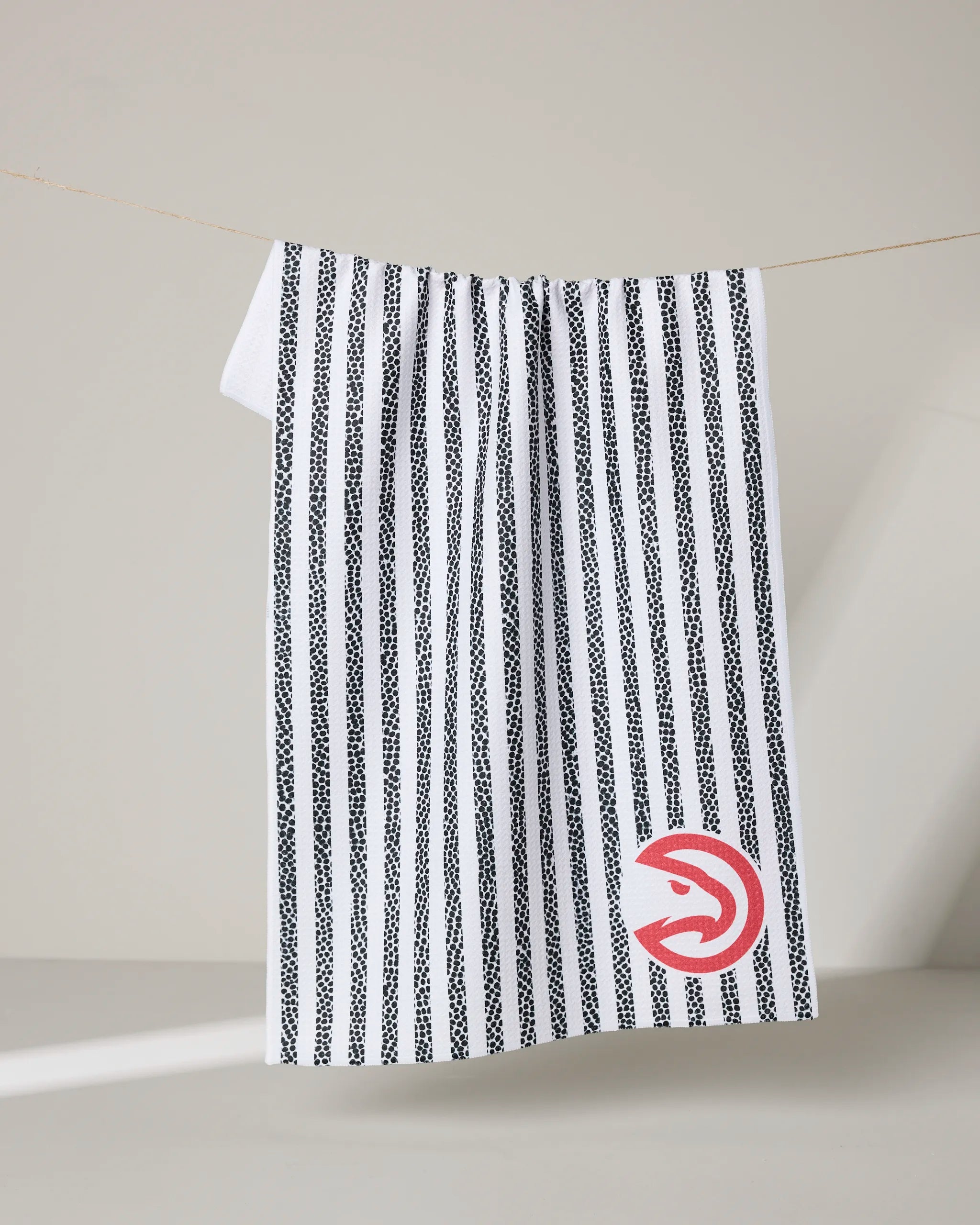 The NBALAB x Geometry Hawks Grip Stripe is a white NBA tea towel with black vertical stripes, hanging on a thin line. Its bottom corner features a red circular Hawks logo, making it an officially licensed sustainable kitchen towel.