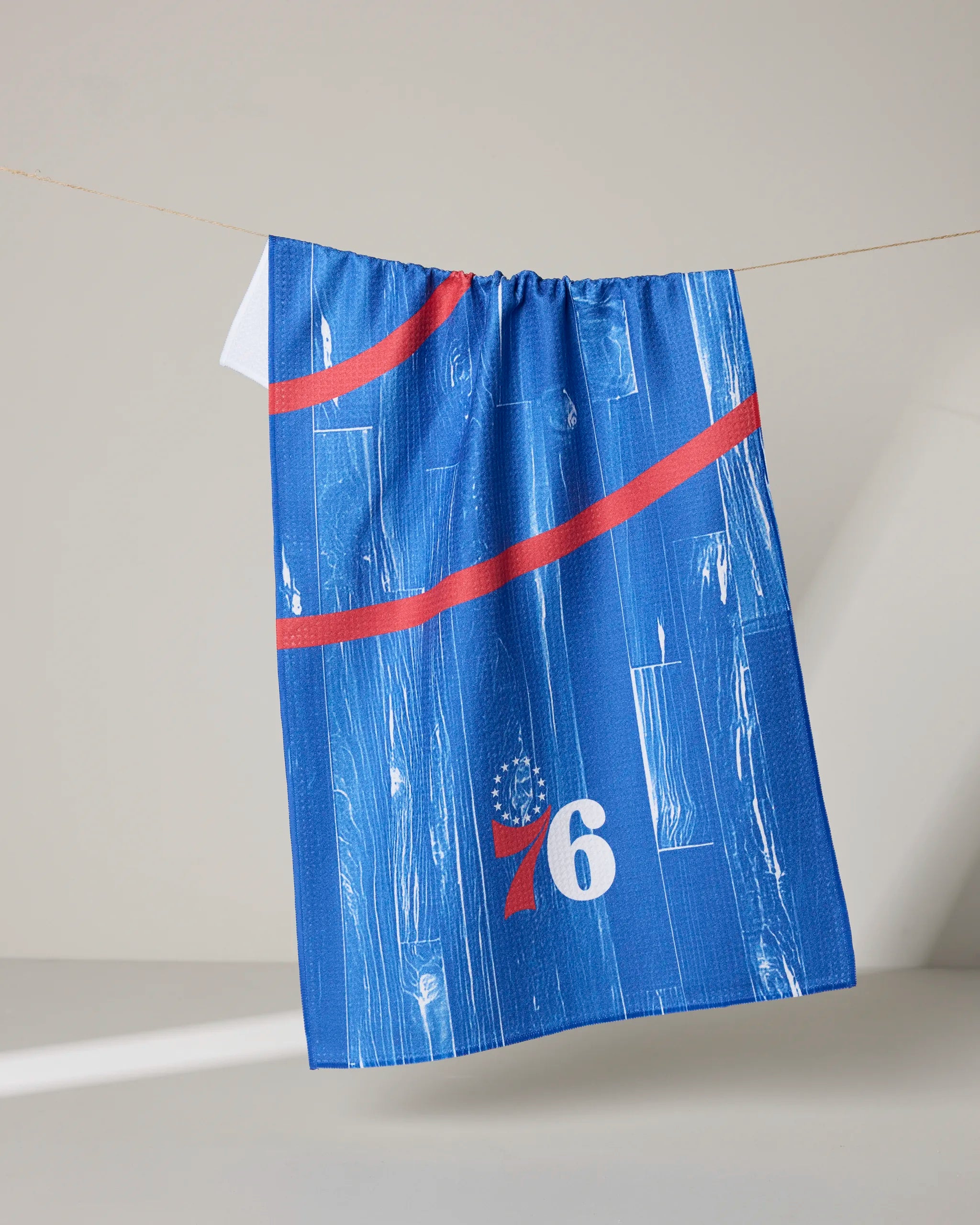 A blue "76ers Zone" jersey by NBALAB x Geometry, with a red stripe, number 76, and white star pattern, hangs on a clothesline resembling an NBA tea towel against a light background.