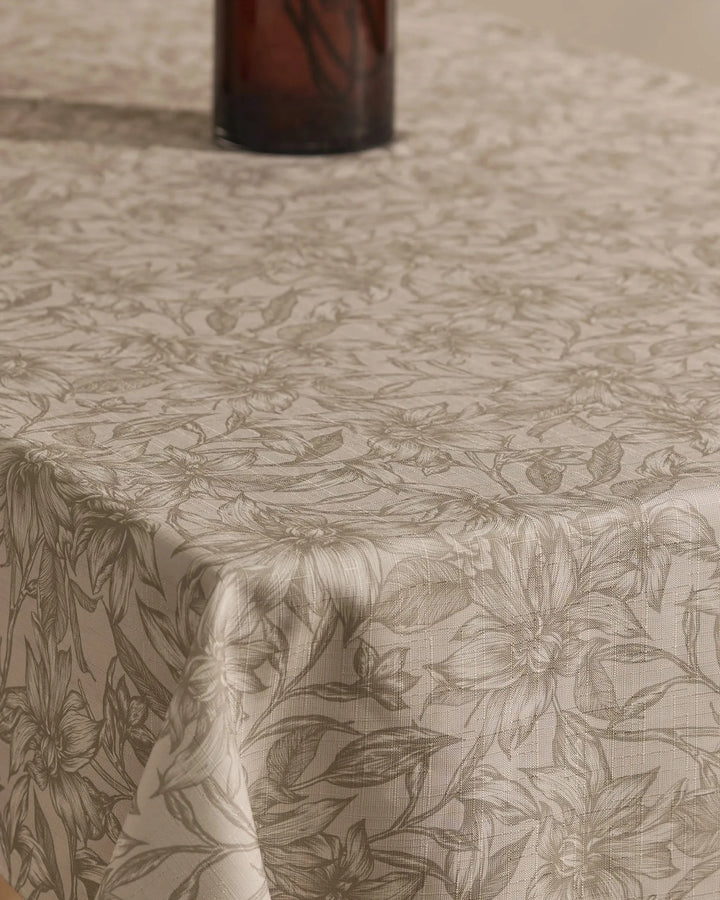 A close-up of the Geometry Alba tablecloth, beige and stain-resistant with a subtle floral pattern, covers the table. The bottom of a dark glass vase appears at the images top.