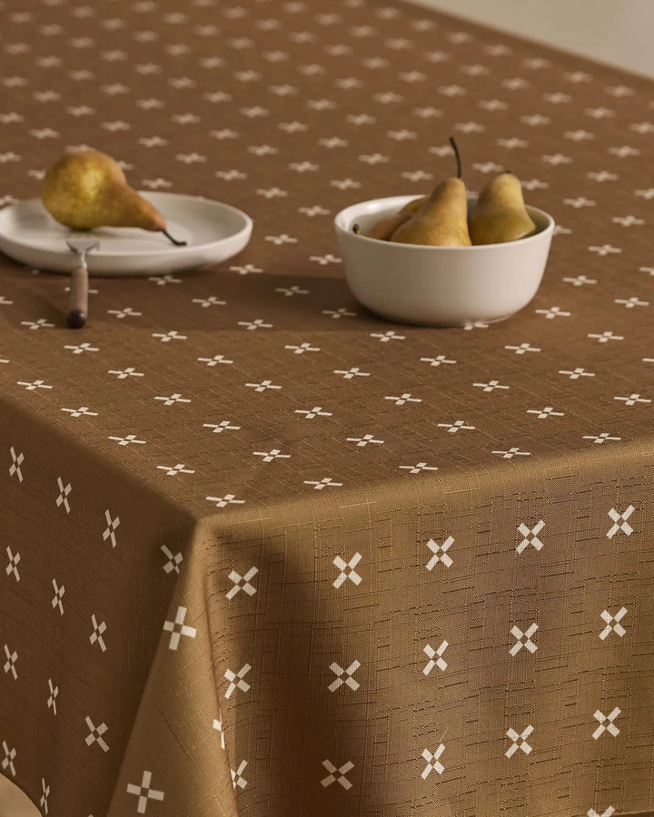 The table is covered with the Amber Stitch tablecloth by Geometry, featuring a white X pattern. A white plate with a pear and a bowl with three pears sit on top, set against a neutral, minimal background.