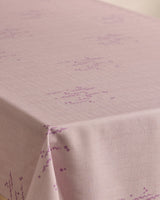 A Bubble Ditzy Dusty Rose tablecloth by Geometry, featuring stain resistance, a subtle grid texture, and small purple geometric patterns, is neatly draped over a table corner.