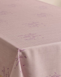 A Bubble Ditzy Dusty Rose tablecloth by Geometry, featuring stain resistance, a subtle grid texture, and small purple geometric patterns, is neatly draped over a table corner.