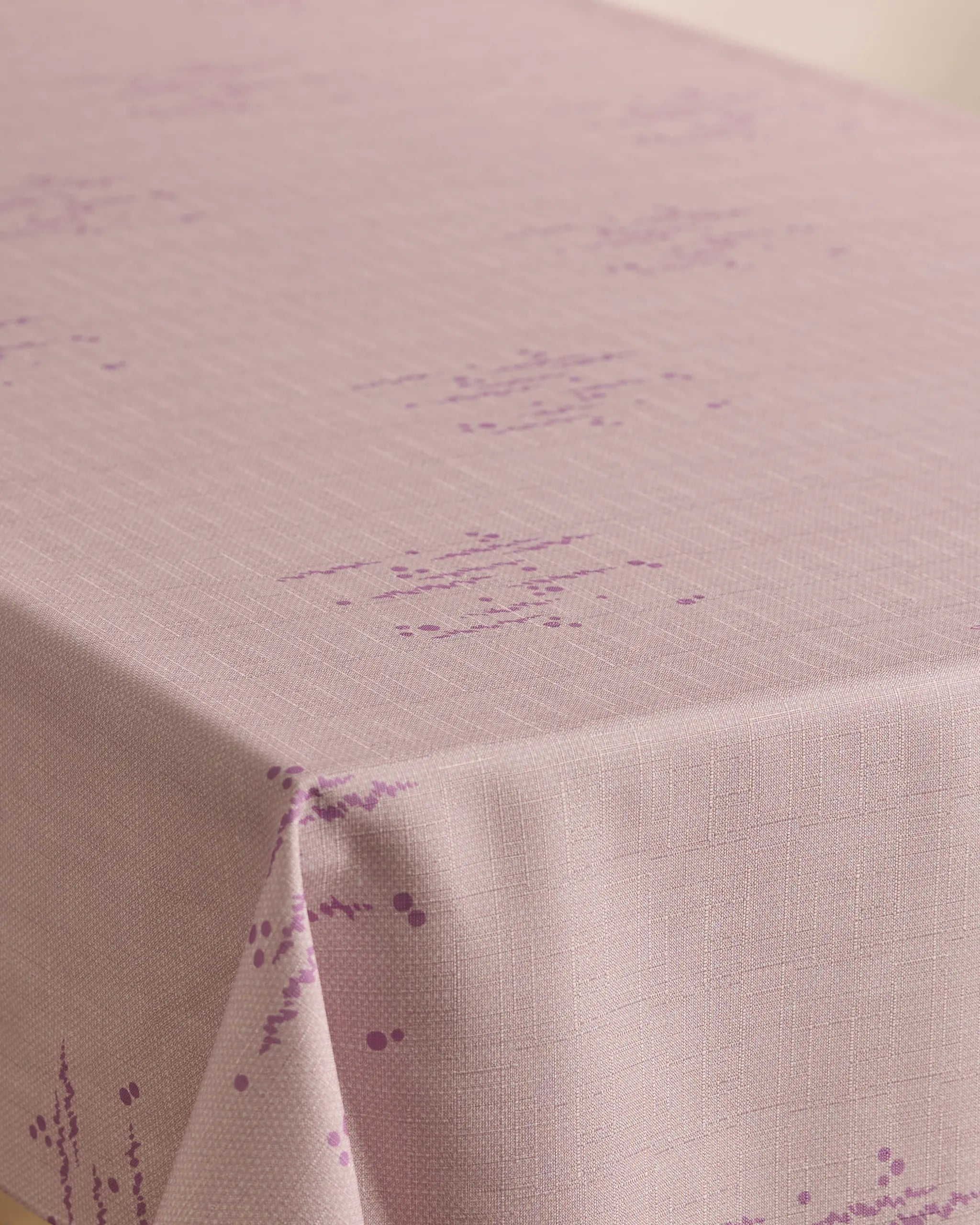 A Bubble Ditzy Dusty Rose tablecloth by Geometry, featuring stain resistance, a subtle grid texture, and small purple geometric patterns, is neatly draped over a table corner.