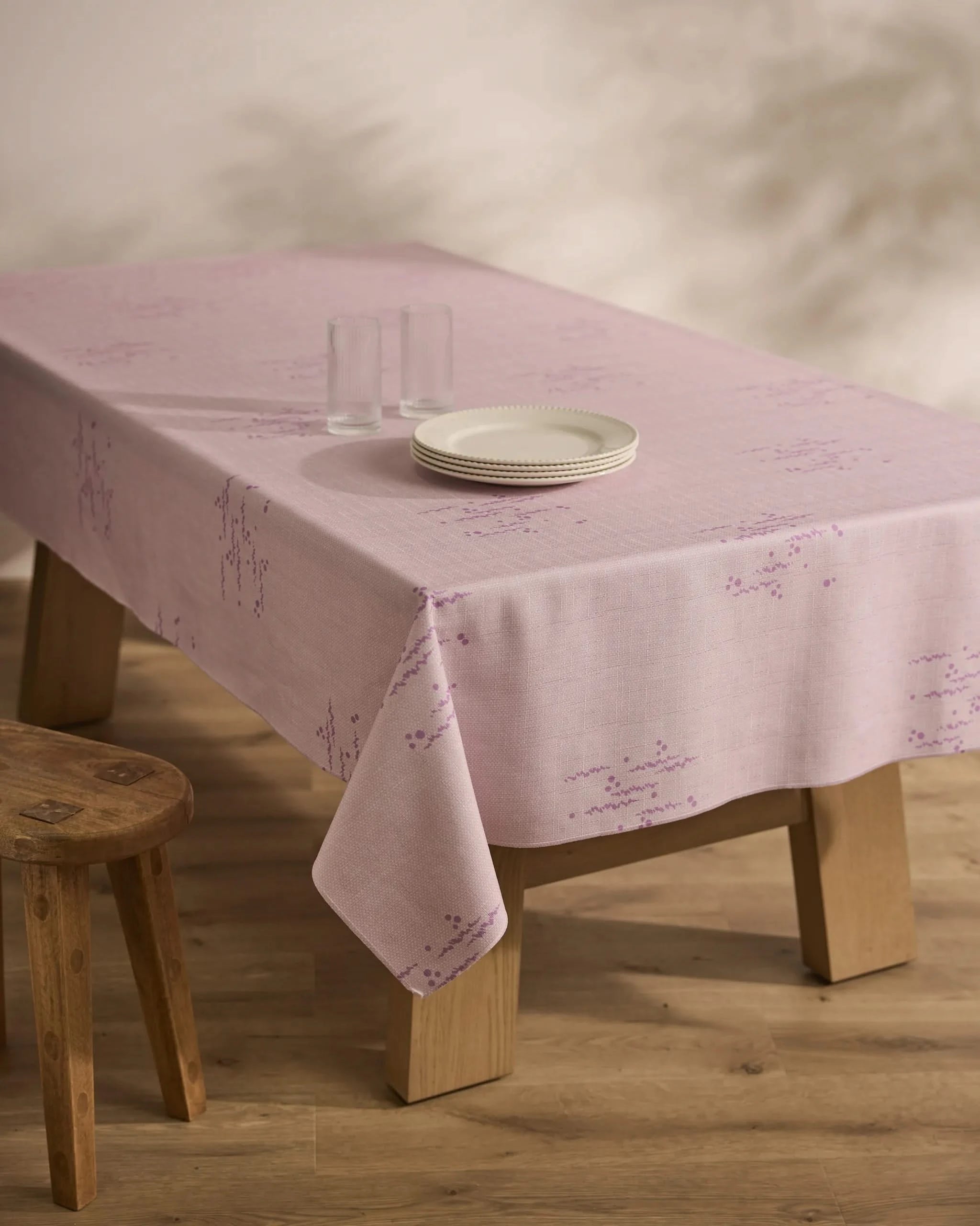 A wooden table draped with the Geometry Bubble Ditzy Dusty Rose stain-resistant tablecloth, featuring subtle patterns, is set with three stacked white plates and two clear glasses beside a basic wooden stool on a wood floor.