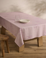 A wooden table draped with the Geometry Bubble Ditzy Dusty Rose stain-resistant tablecloth, featuring subtle patterns, is set with three stacked white plates and two clear glasses beside a basic wooden stool on a wood floor.