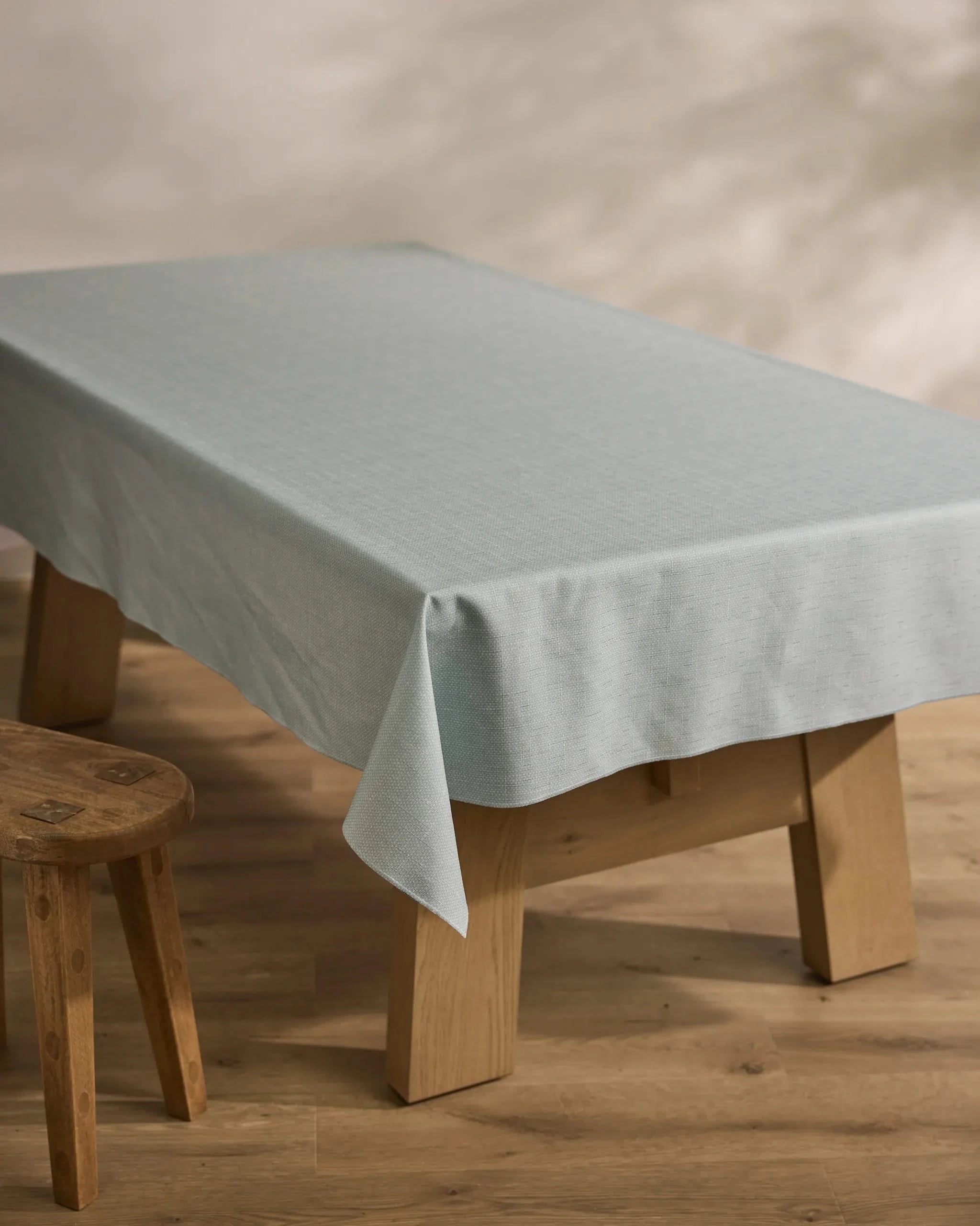 A wooden table draped with the Geometry Chambray Light Blue sustainable tablecloth stands on a wooden floor beside a small wooden stool, with a softly blurred neutral-toned background.