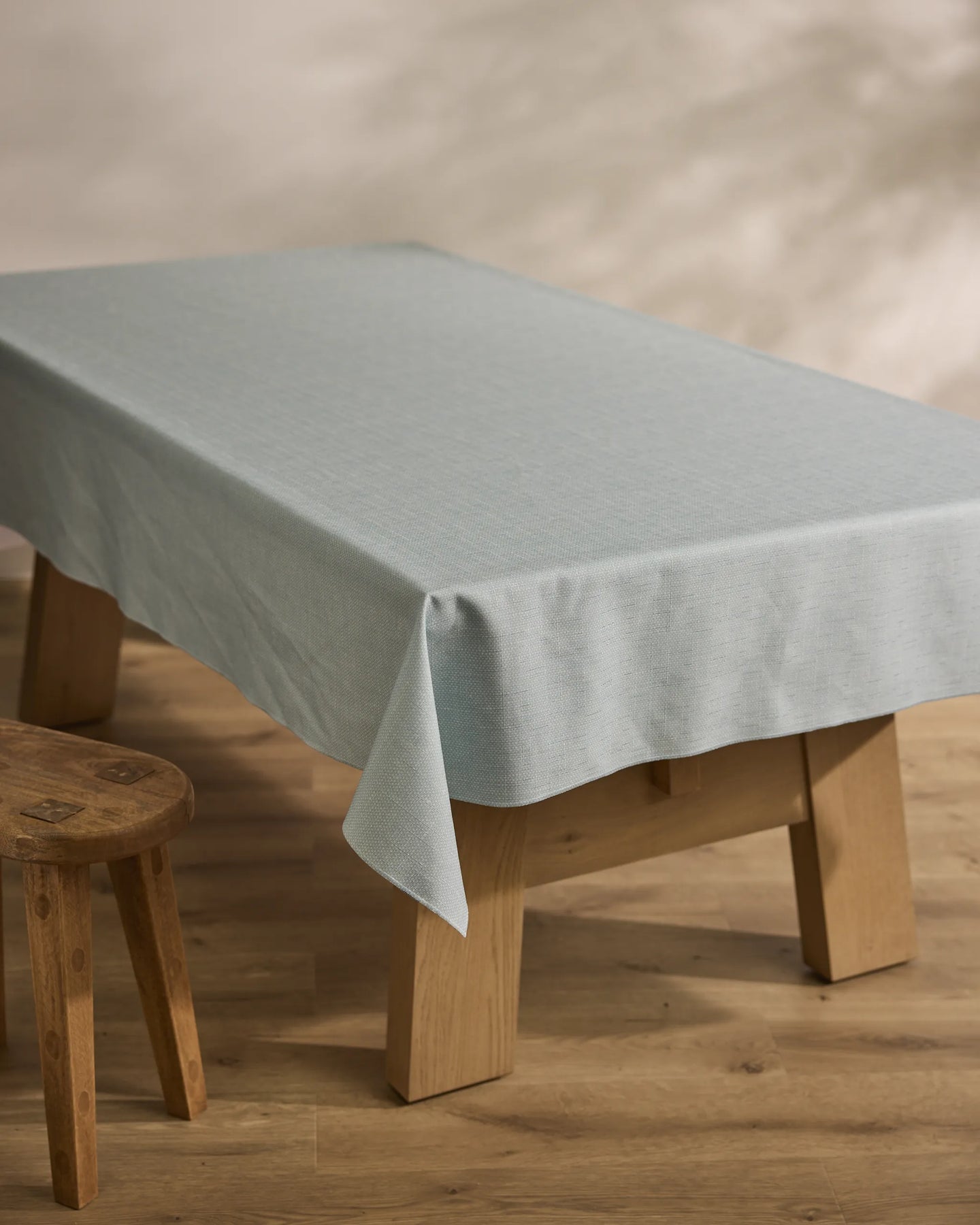 A wooden table draped with the Geometry Chambray Light Blue sustainable tablecloth stands on a wooden floor beside a small wooden stool, with a softly blurred neutral-toned background.