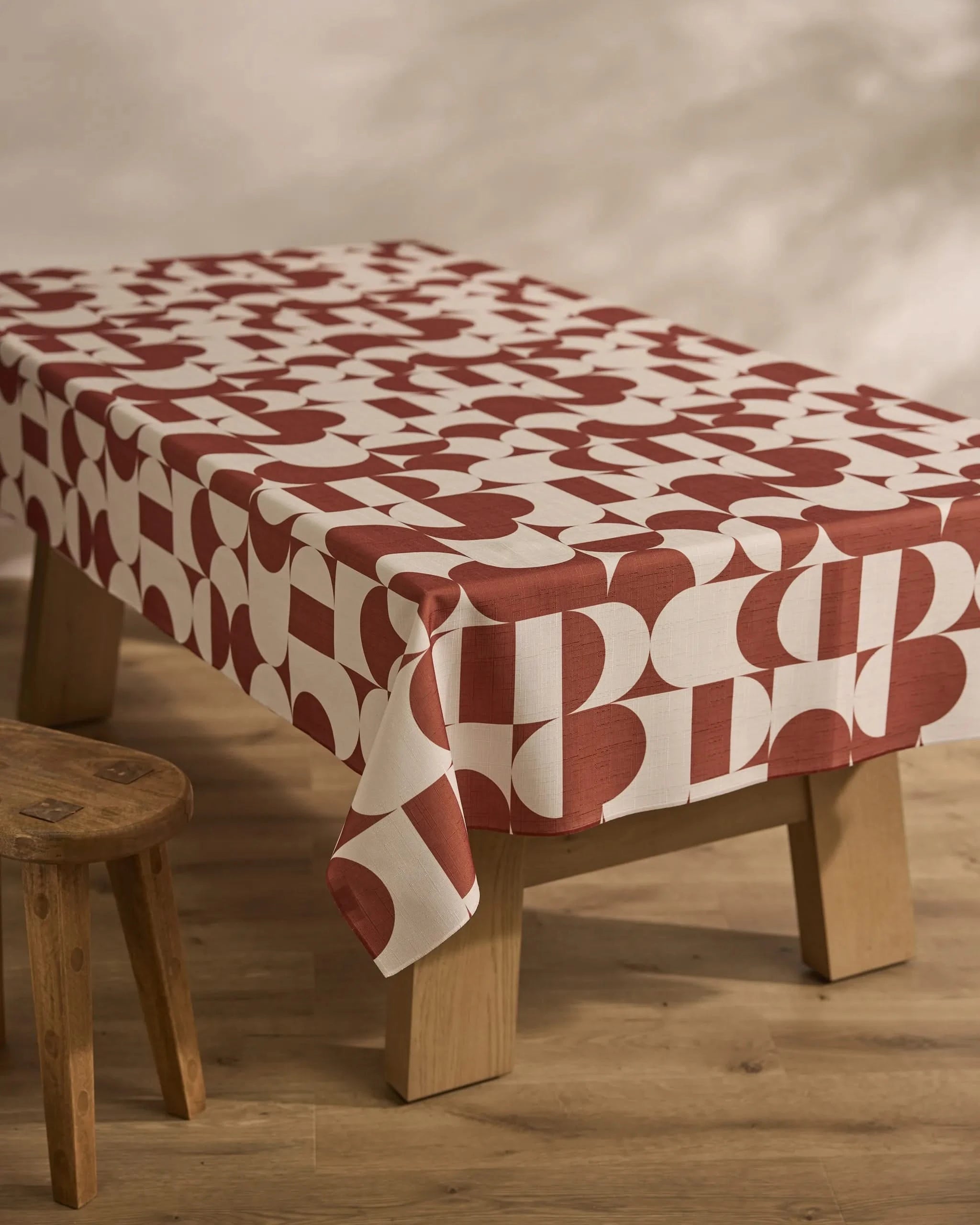 A wooden table features the Geometry Circle Playground sustainable tablecloth in a red and white geometric pattern with bold circles and rectangles, set on a wooden floor beside a small wooden stool.