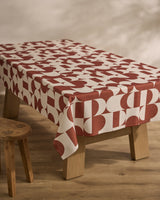 A wooden table features the Geometry Circle Playground sustainable tablecloth in a red and white geometric pattern with bold circles and rectangles, set on a wooden floor beside a small wooden stool.