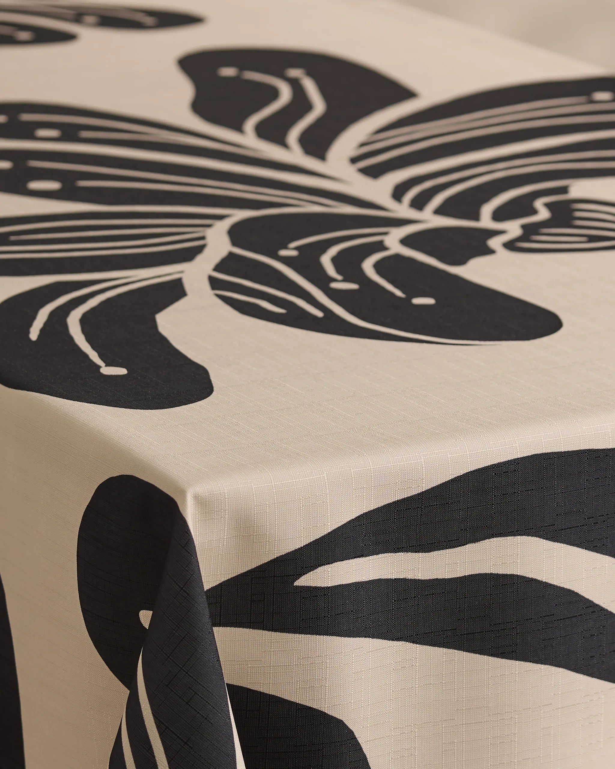 A close-up of a table featuring Geometrys Giant Lush Floral tablecloth, cream-colored and stain-resistant, decorated with a bold abstract black leaf design that extends over the edge.
