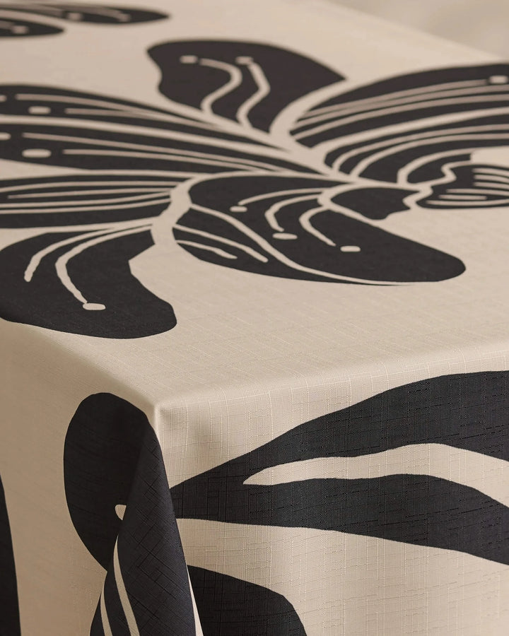 A close-up of a table featuring Geometrys Giant Lush Floral tablecloth, cream-colored and stain-resistant, decorated with a bold abstract black leaf design that extends over the edge.