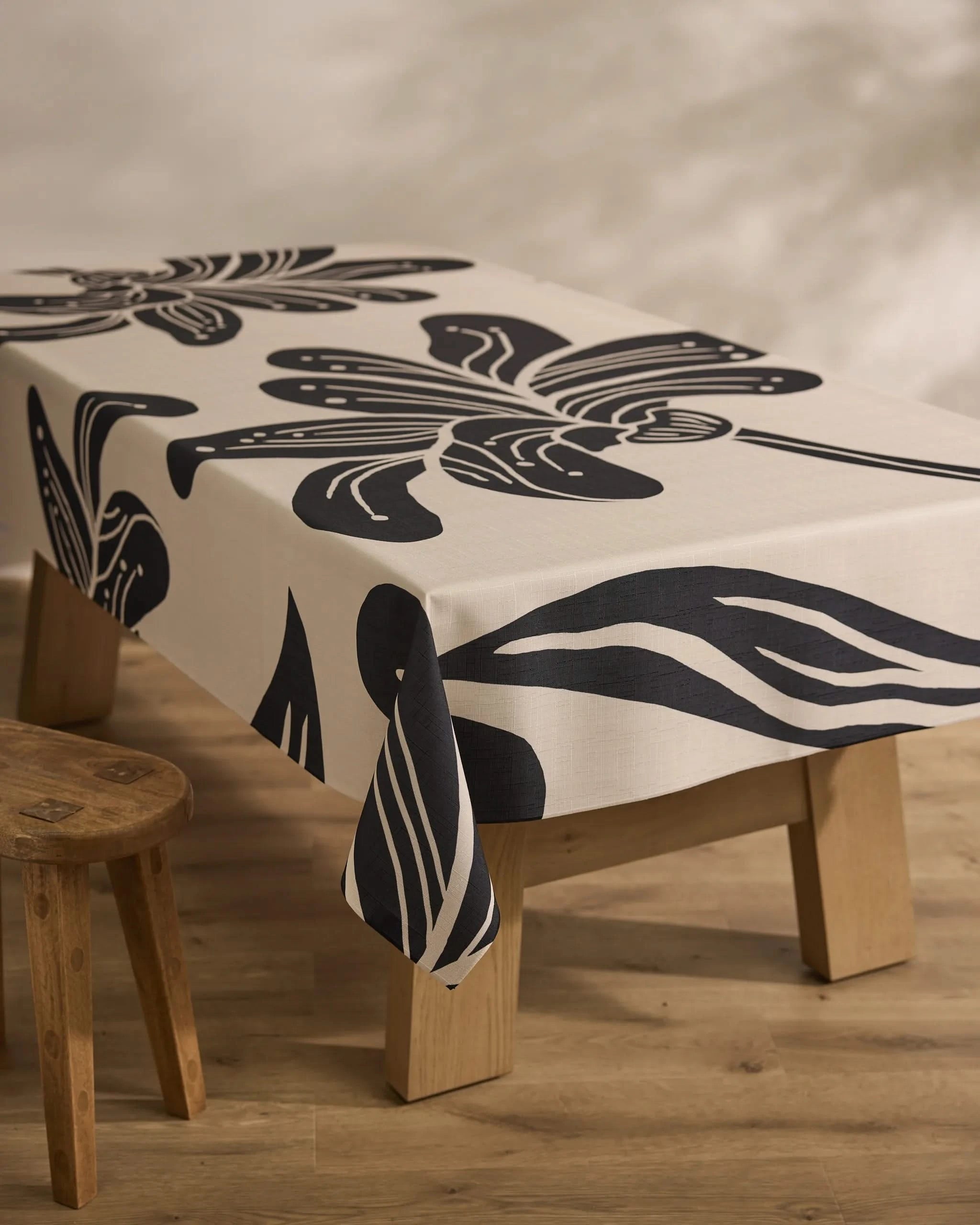 A rectangular wooden table is draped with Geometry’s Giant Lush Floral cream tablecloth, showcasing bold black abstract florals. A rustic stool sits beside the table on a light wood floor.