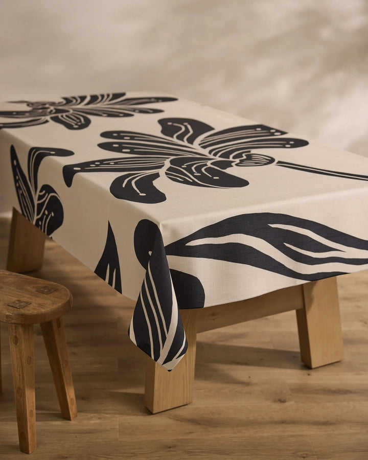 A rectangular wooden table is draped with Geometry’s Giant Lush Floral cream tablecloth, showcasing bold black abstract florals. A rustic stool sits beside the table on a light wood floor.