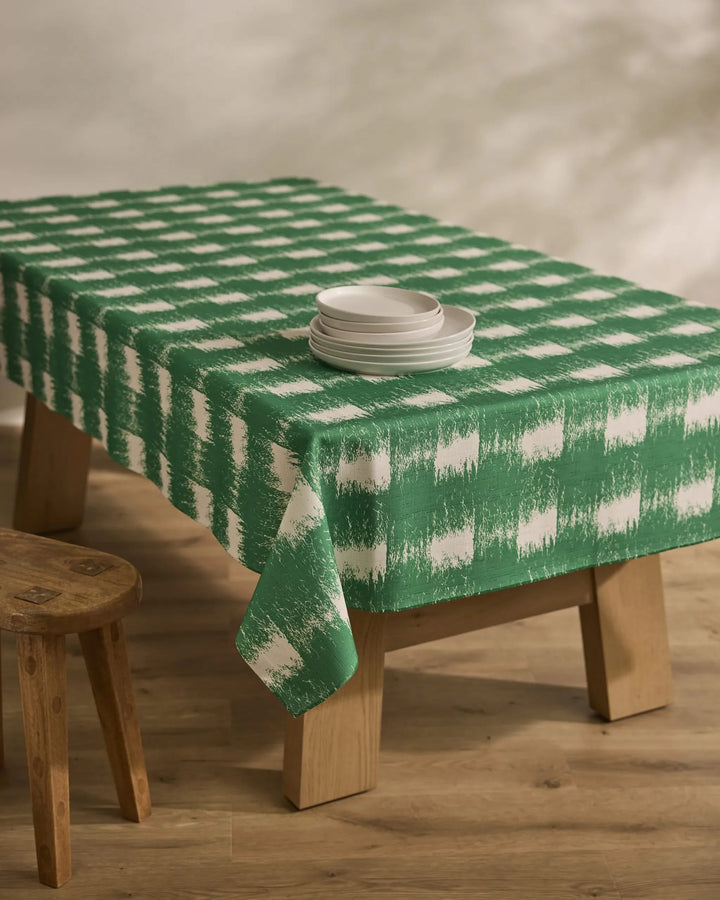 A wooden table is draped with the Geometry Hairy Check Green tablecloth and set with stacked white plates. A wooden stool sits nearby on a light wood floor, all lit by soft, natural light.