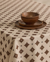 The Heritage Stamp tablecloth by Geometry, in beige with a brown geometric pattern, covers the table. Two wooden plates are stacked and two wooden bowls are nested, creating a simple, rustic setting.