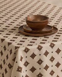 The Heritage Stamp tablecloth by Geometry, in beige with a brown geometric pattern, covers the table. Two wooden plates are stacked and two wooden bowls are nested, creating a simple, rustic setting.