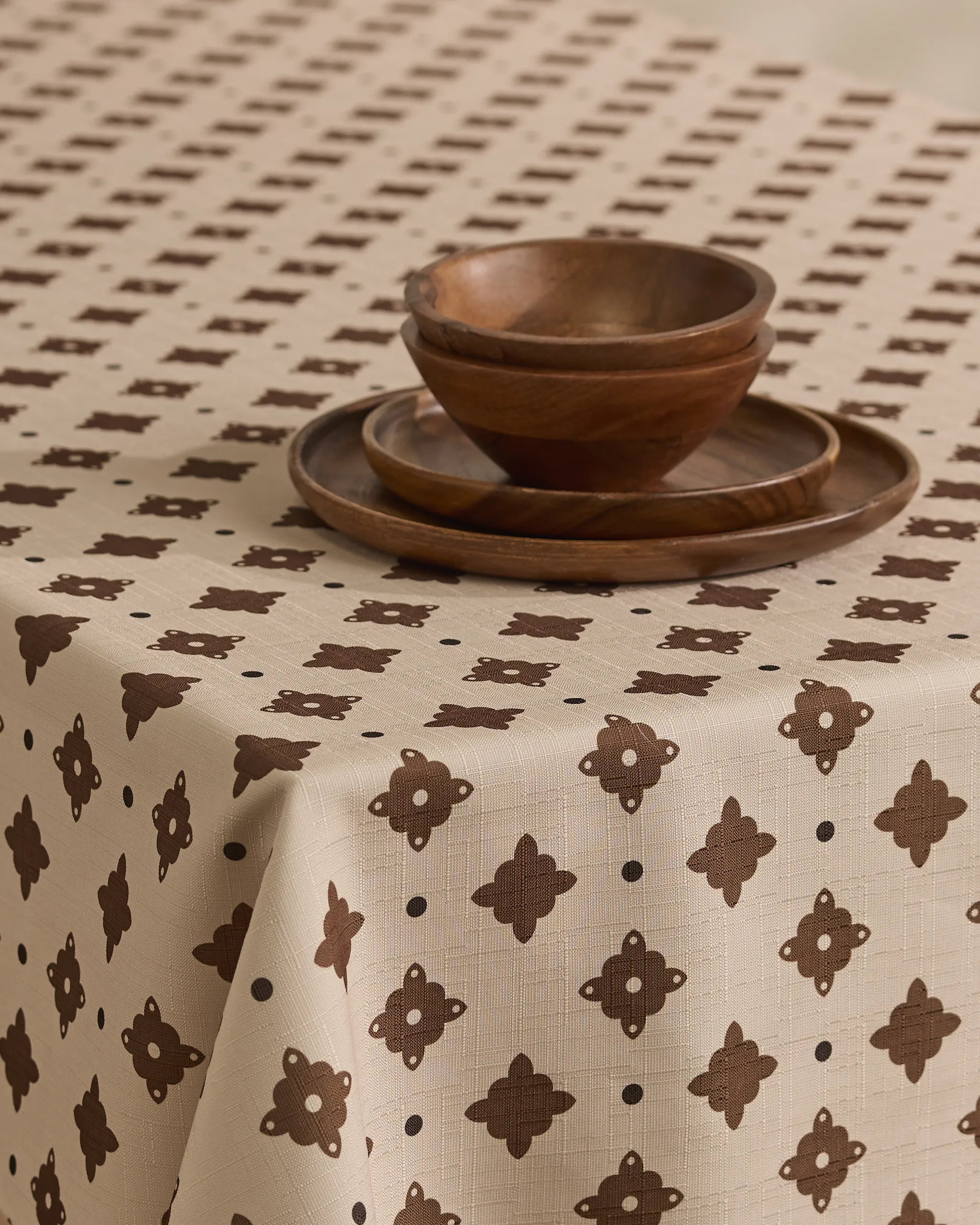 The Heritage Stamp tablecloth by Geometry, in beige with a brown geometric pattern, covers the table. Two wooden plates are stacked and two wooden bowls are nested, creating a simple, rustic setting.