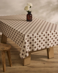 The Geometry Heritage Stamp table features an eco-friendly beige and brown patterned tablecloth, a small brown glass vase with white flowers, and is paired with a wooden stool on a light wood floor.