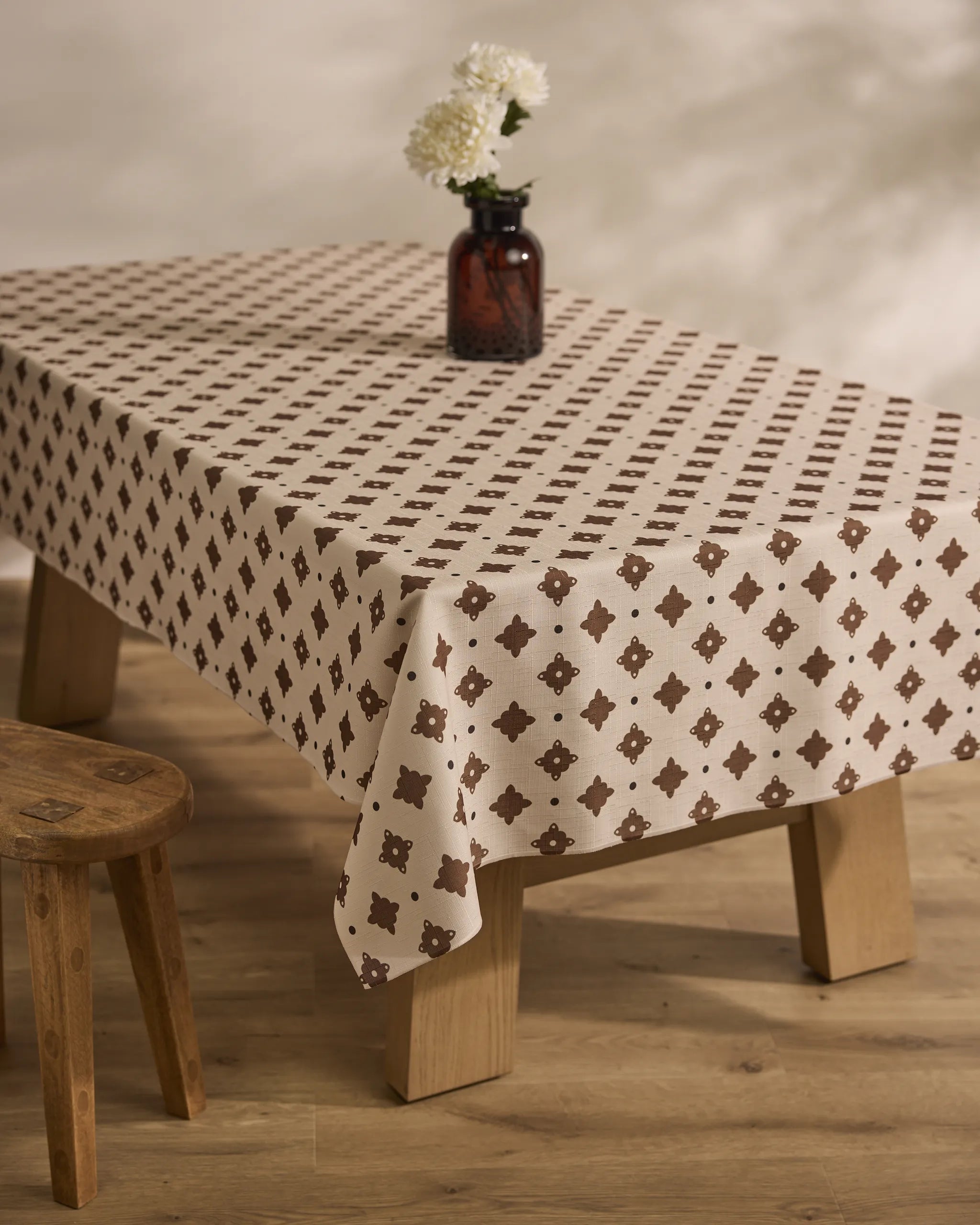 The Geometry Heritage Stamp table features an eco-friendly beige and brown patterned tablecloth, a small brown glass vase with white flowers, and is paired with a wooden stool on a light wood floor.