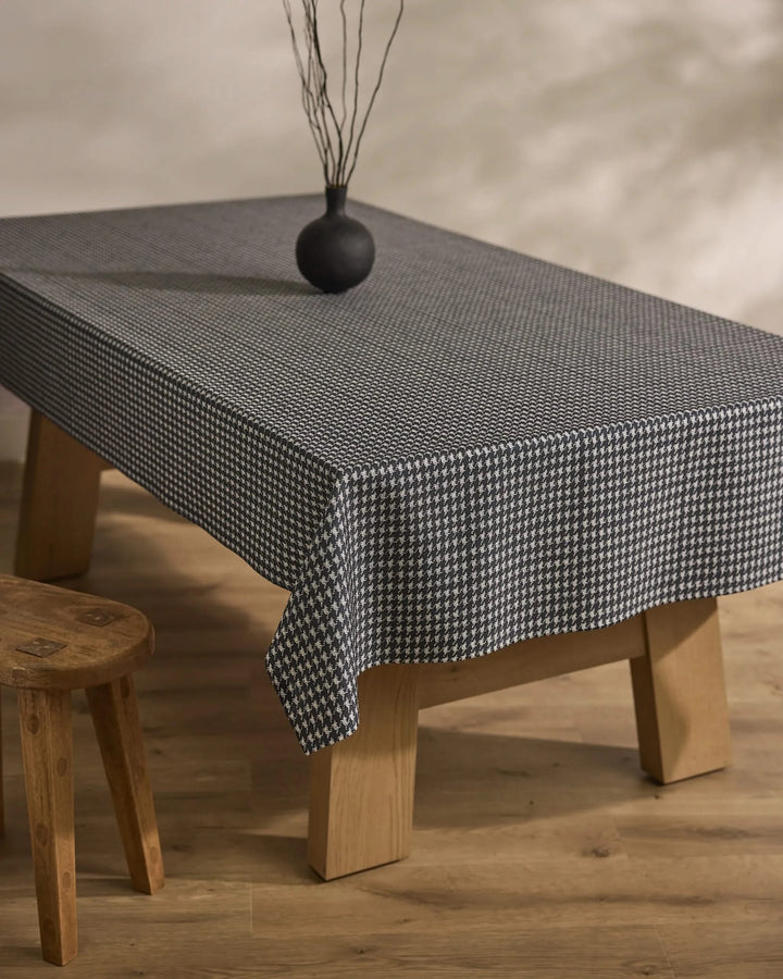 A rectangular wooden table is draped with Geometry’s Houndstooth Black tablecloth, topped with a small round black vase holding dry branches. A wooden stool sits beside the table on a light wooden floor.
