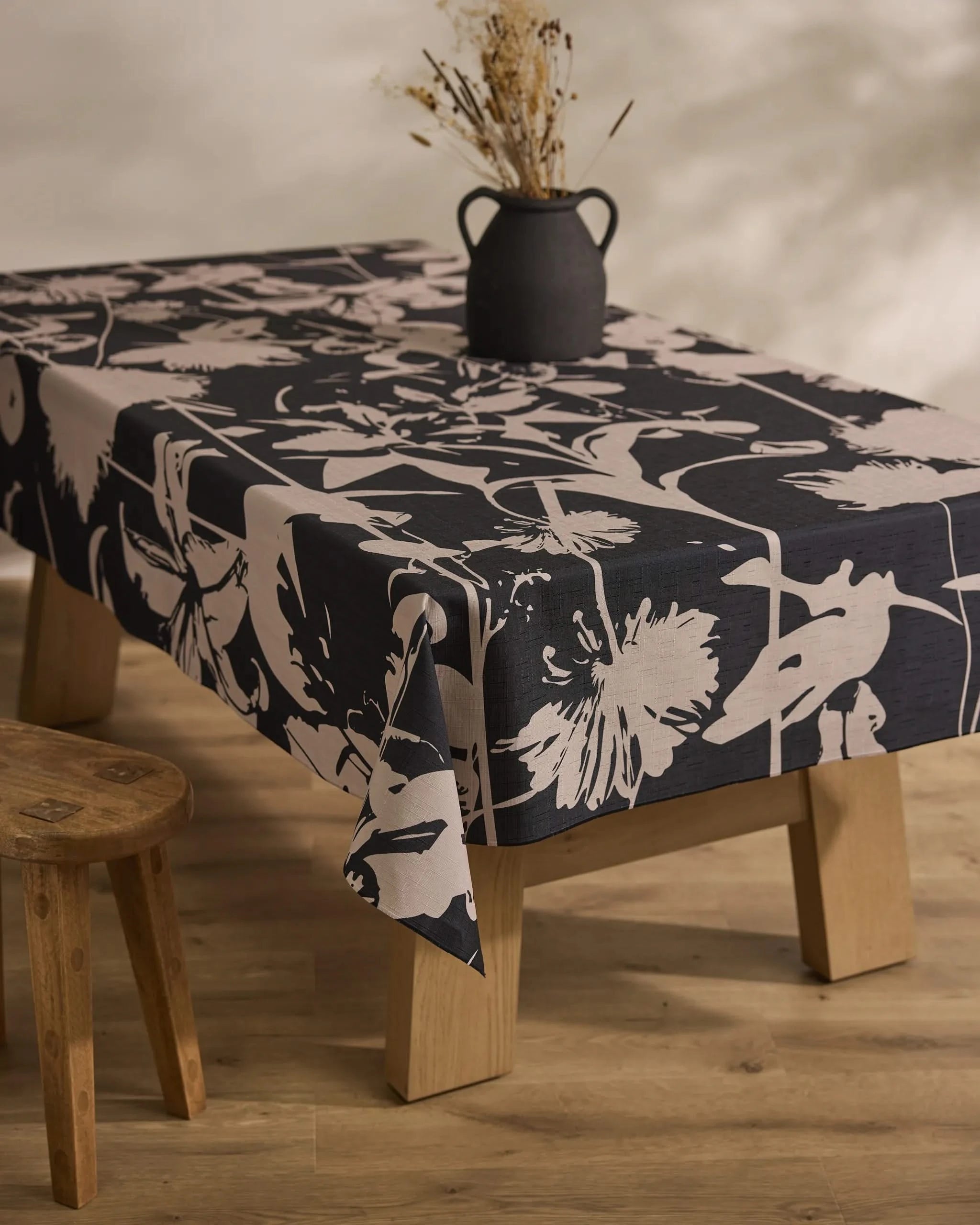 A wooden table draped with the Inverted Floral Black stain-resistant tablecloth from Geometry, topped with a rustic black vase of dried flowers and paired with a simple wooden stool, rests on a light wood floor.