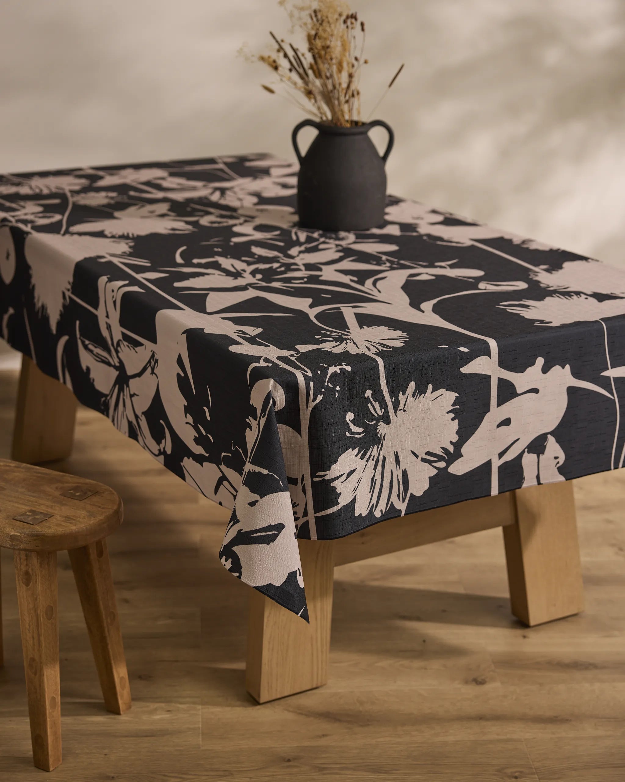 A wooden table draped with the Inverted Floral Black stain-resistant tablecloth from Geometry, topped with a rustic black vase of dried flowers and paired with a simple wooden stool, rests on a light wood floor.