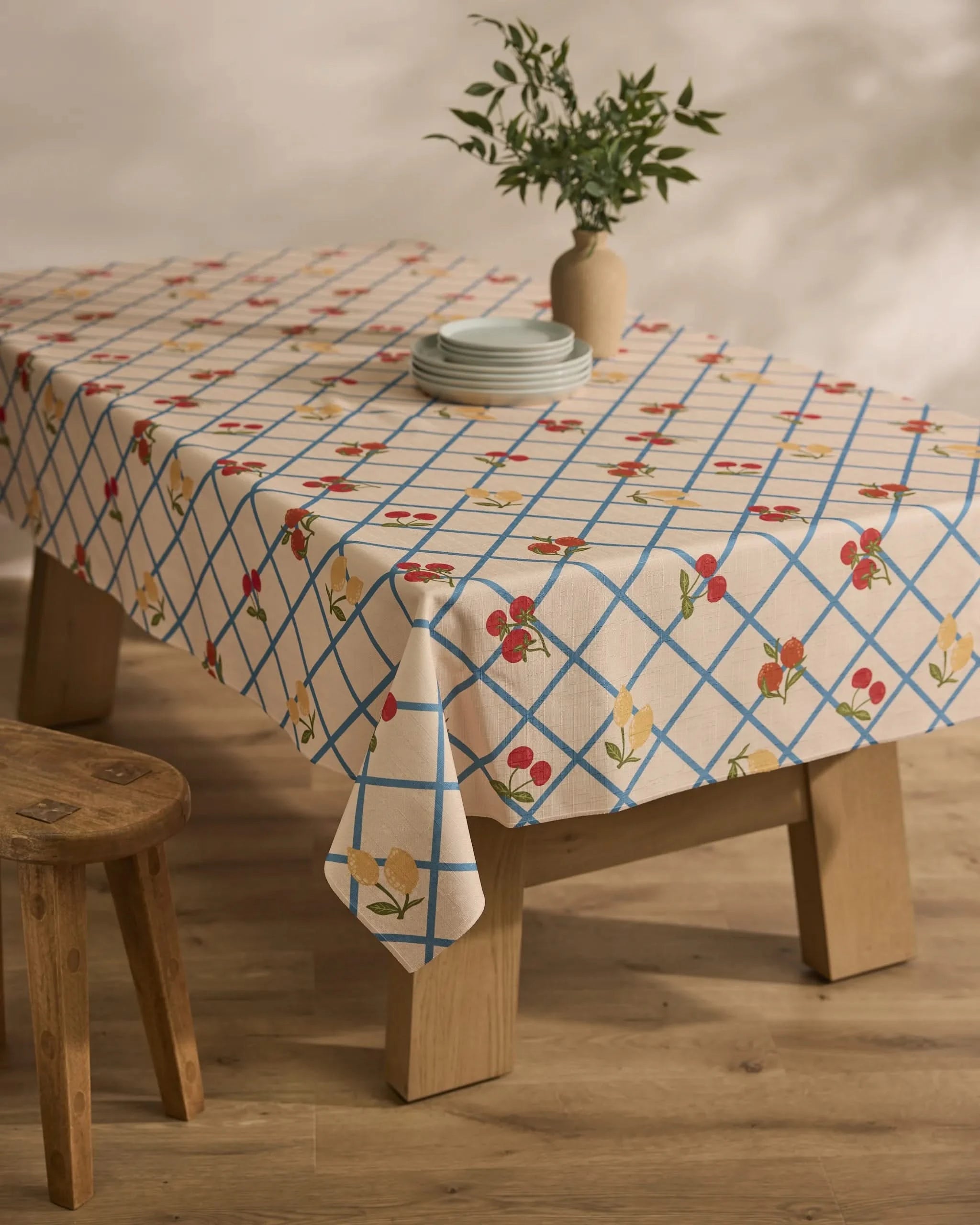 A wooden table is draped with the Geometry Italian Summer Grid tablecloth, showcasing blue grid lines and red and yellow flowers, set with plates and a vase of green foliage. Nearby, a wooden stool rests on a wooden floor.