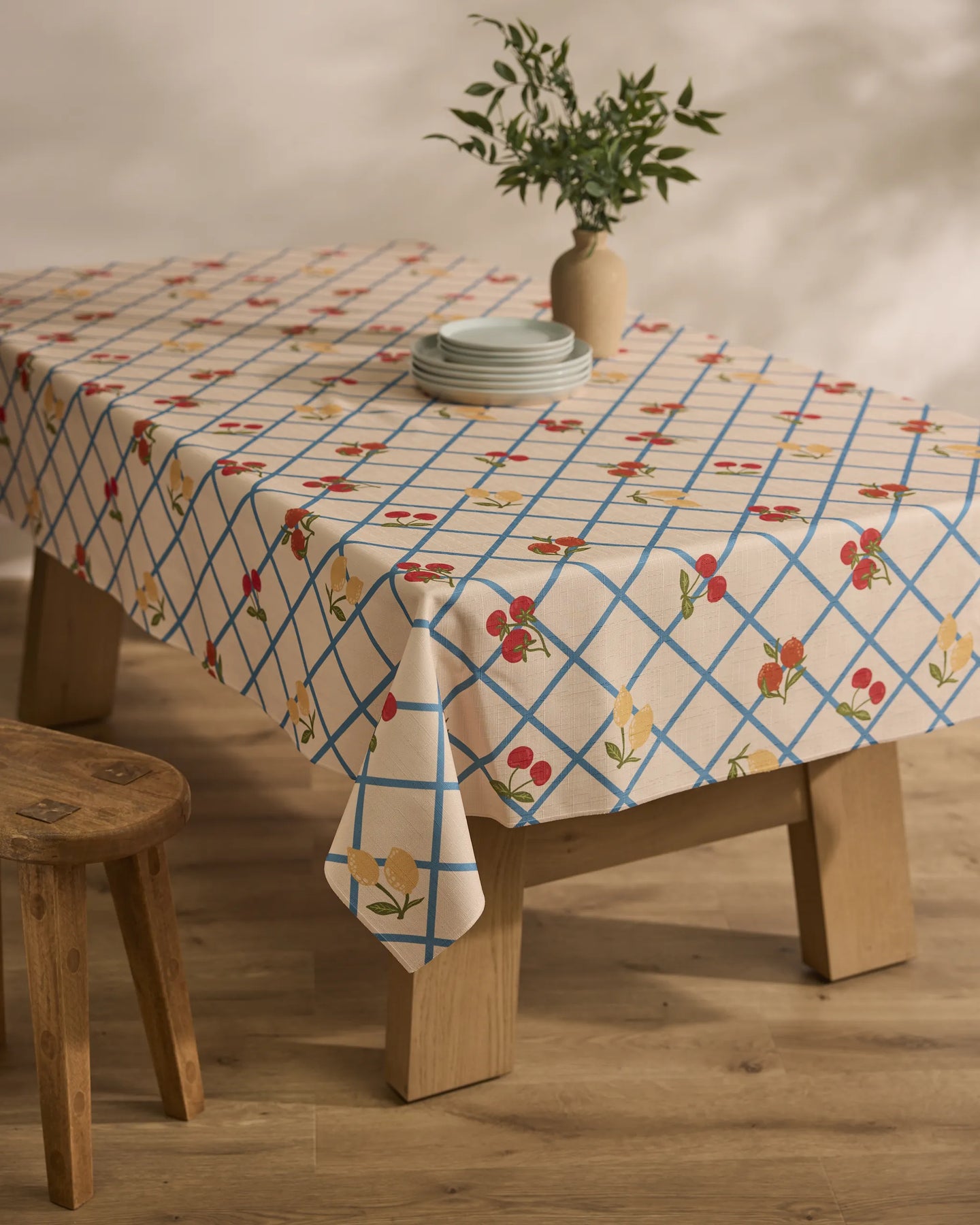 A wooden table is draped with the Geometry Italian Summer Grid tablecloth, showcasing blue grid lines and red and yellow flowers, set with plates and a vase of green foliage. Nearby, a wooden stool rests on a wooden floor.