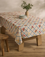 A wooden table is draped with the Geometry Italian Summer Grid tablecloth, showcasing blue grid lines and red and yellow flowers, set with plates and a vase of green foliage. Nearby, a wooden stool rests on a wooden floor.