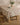 A wooden table is draped with the Geometry Italian Summer Grid tablecloth, showcasing blue grid lines and red and yellow flowers, set with plates and a vase of green foliage. Nearby, a wooden stool rests on a wooden floor.