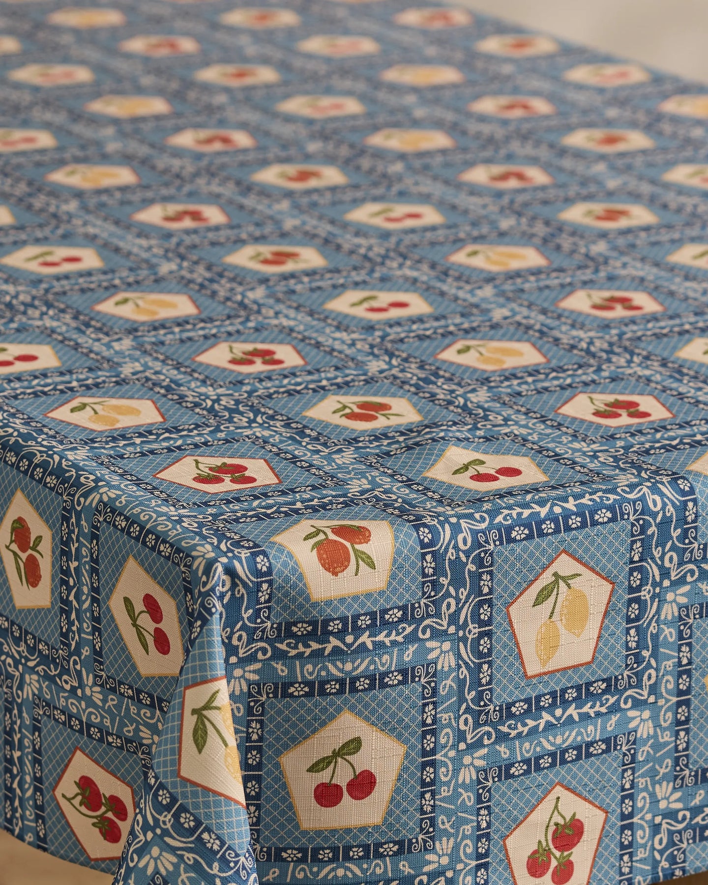 A rectangular table is covered with Geometry’s Italian Tiles tablecloth, which has a blue background, repeating white designs, and fruit illustrations like cherries, lemons, and strawberries inside geometric shapes.