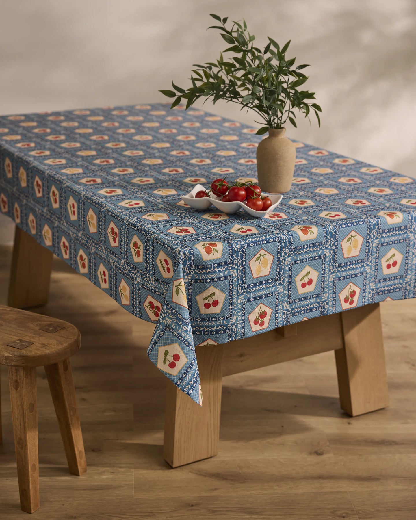 A wooden table is topped with Geometry’s Italian Tiles blue patterned, stain-resistant tablecloth adorned in red and yellow. A beige vase with green leafy branches and a plate of tomatoes sit atop. Nearby is a wooden stool on a light wood floor.