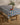 A wooden table is topped with Geometry’s Italian Tiles blue patterned, stain-resistant tablecloth adorned in red and yellow. A beige vase with green leafy branches and a plate of tomatoes sit atop. Nearby is a wooden stool on a light wood floor.