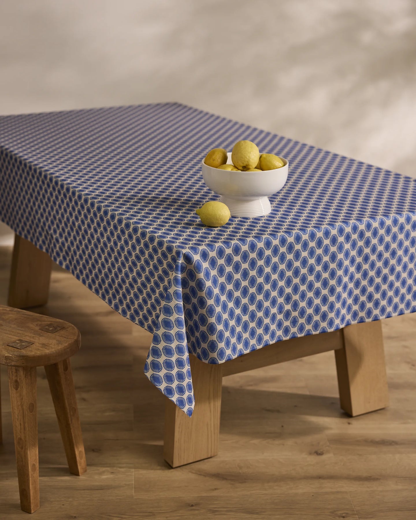 A Lagoon tablecloth by Geometry covers a wooden table that displays a white bowl of lemons with two beside it; a wooden stool is nearby on the light wood floor.